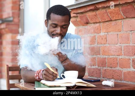 Junger afroamerikanischer Geschäftsmann, der elektronische Zigaretten raucht und sich Notizen in Notizbüchern macht, während er im Café im Freien am Tisch sitzt Stockfoto