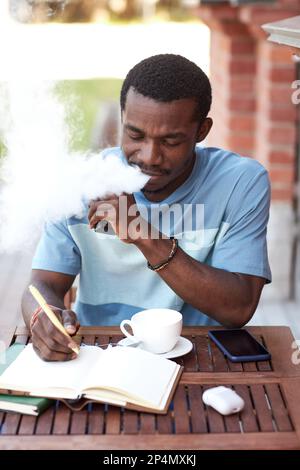 Junger afroamerikanischer Geschäftsmann, der Vape Pen raucht, sitzt am Tisch im Café im Freien und macht sich Notizen in Notizbuch, während er seine Arbeit organisiert Stockfoto