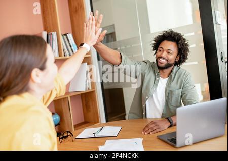 Zwei verschiedene Mitarbeiter in eleganter Freizeitkleidung geben sich im modernen Tagungsraum gegenseitig fünf Punkte und feiern die erfolgreiche Zusammenarbeit. Die Kollegen unterstützen sich gegenseitig Stockfoto