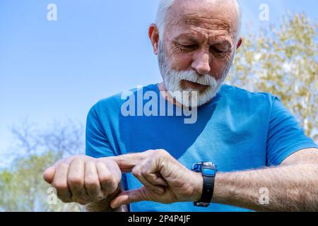 Porträt eines Seniorenmannes, der auf seine Uhr schaut.Seniorensportler überwacht seine Herzfrequenz und seinen Blutdruck mit der Smartwatch am Handgelenk. Stockfoto