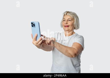 Graue ältere Frau, die während der Selfie-Aufnahme auf dem Mobiltelefon auf weißem Hintergrund isoliert gestikuliert Stockfoto