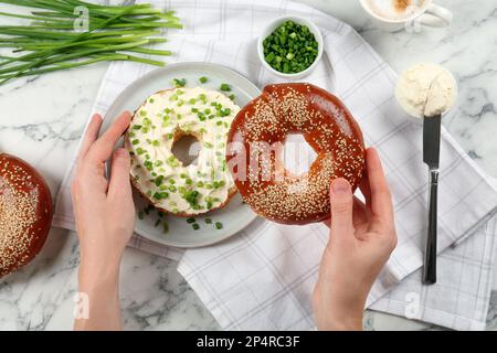 Frau, die Bagel mit Frischkäse und grünen Zwiebeln am weißen Marmortisch macht, Draufsicht Stockfoto