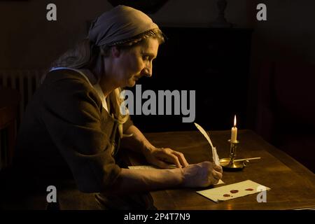 Eine Frau in mittelalterlicher Kleidung, die in einem dunklen Raum sitzt und einen Brief mit Federdecke schreibt Stockfoto