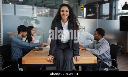 Zufriedene lächelnde hispanische Geschäftsfrau, leitende Mitarbeiterin, die im Vorstandsbüro auf einem Tisch sitzt, mit Kollegen aus mehreren ethnischen Gruppen Stockfoto
