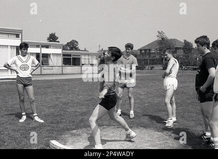 1989, historischer Sporttag der Sekundarschule, ein Teenager, der den Shot Put macht, beobachtet von anderen männlichen Schülern, Sleaford, Lincs, England, Großbritannien. Der Shot Put ist ein athletisches Ereignis, bei dem ein schwerer Kugelball, so weit wie möglich, aus einem kleinen Kreis in England, in Großbritannien, geworfen wird Stockfoto