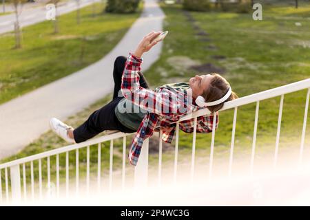 Eine Frau, die auf dem Geländer liegt und draußen im Park Musik hört. Stockfoto