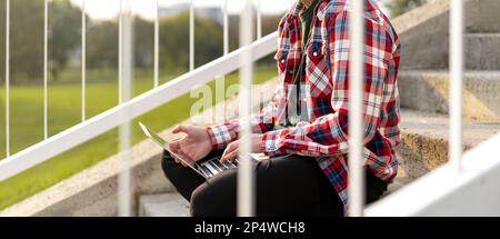 Nicht binäre Person, die auf den Stufen im Freien im Park sitzt und an einem Laptop arbeitet. Stockfoto