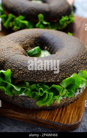 Sandwiches mit schwarzen Bagels und Gurken, Salat und Schinken auf Holzbrett und dunkler Hintergrundfarbe, leckerer Snack oder Frühstück Stockfoto