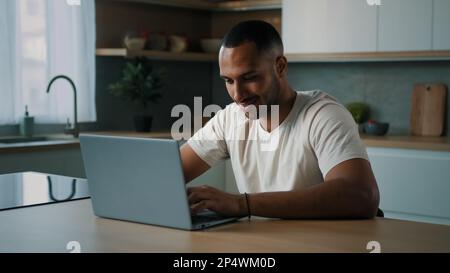 Afrikanischer Millennials Multirassist, amerikanischer Freiberufler mit männlichen Benutzern, der am Küchenschreibtisch sitzt und einen Laptop verwendet, der entfernt mit einer Computer-App arbeitet Stockfoto
