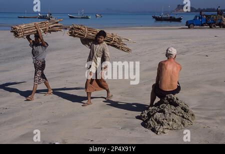 24.02.2008, Thandwe, Rakhine State, Myanmar, Asien - zwei Einheimische mit Feuerholz laufen vorbei an einem Touristen am Ngapali Beach, der auf einem Felsen sitzt. Stockfoto