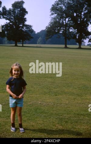 Ein kleines 5-jähriges Mädchen in hellblauen Shorts und dunkelblauem T-Shirt, das in den 1970er Jahren im Park in den Vororten von London, Großbritannien, stand Stockfoto