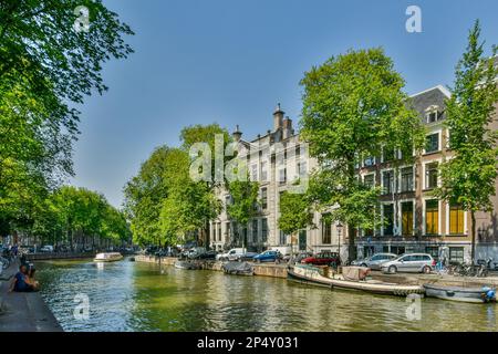 Amsterdam, Niederlande - 10. April 2021: Einige Gebäude und Boote auf dem Wasser vor ihnen, mit Bäumen an den Seiten des Kanals Stockfoto