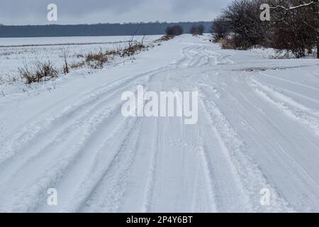 Verschneite Straße in einem Feld, das zum Kiefernwald führt. Winterstraße ins Nirgendwo an sonnigen Tagen, schneebedeckte frische Autorasse. Auto zeichnet in einem tiefen Schnee von abgelegenen r nach Stockfoto