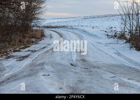 Verschneite Straße in einem Feld, das zum Kiefernwald führt. Winterstraße ins Nirgendwo an sonnigen Tagen, schneebedeckte frische Autorasse. Auto zeichnet in einem tiefen Schnee von abgelegenen r nach Stockfoto