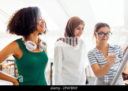 Brainstorming mit drei Teenagern. Teenager verschiedener Nationalitäten und Religionen lösen Probleme gemeinsam. Das weiße Mädchen schreibt mit Markern an die Tafel. Stockfoto