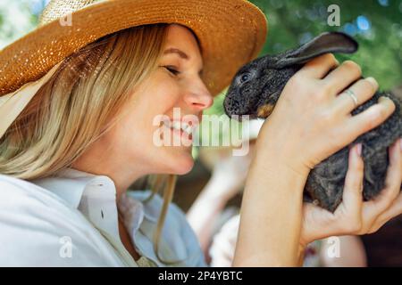 Nahaufnahme einer jungen, schönen Frau mit einem kleinen schwarzen Kaninchen. Ein hübsches, weißes Mädchen mit Strohhut und weißem Hemd lächelt und hält ein süßes Häschen in der Hand Stockfoto