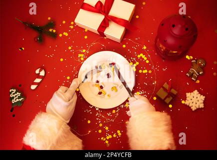 Weihnachtsmann mit Teller mit Tabletten, Pillen und Kapseln. Hände in weißen Handschuhen halten Messer und Gabel. Geschenkschachteln, weihnachtskekse, Laterne und Tannen-BH Stockfoto