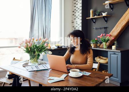 Glückliche junge afro-amerikanische Frau, die zu Hause sitzt, ihren Laptop benutzt und auf der Tastatur tippt. Lächelnde Studentin oder Freiberuflerin, die auf einen Computerbildschirm schaut. Stockfoto