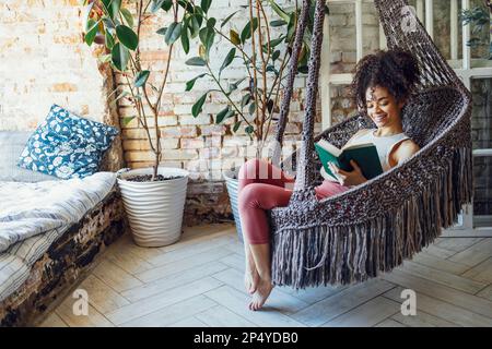 Ein junges, wunderschönes afro-amerikanisches Mädchen liest in einem Korbstuhl. Dunkelhäutiger Teenager in legerer Kleidung ruht zu Hause aus. Weibliches Teenager-Lächeln Stockfoto