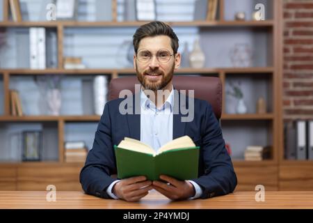 Porträt eines Mannes, eines Pastors einer Kirche, einer religiösen Gemeinschaft verschiedener Konfessionen, der seine Predigt online hält. Im Büro sitzen und in die Kamera schauen, ein heiliges Buch in der Hand halten. Stockfoto