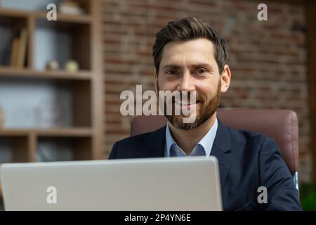 Nahaufnahme eines erfolgreichen, erwachsenen Geschäftsmanns im Heimbüro, bärtigen Mannes bei der Arbeit mit einem lächelnden Laptop und einem Blick auf die Kamera, der mit der Fernarbeit zufrieden ist. Stockfoto