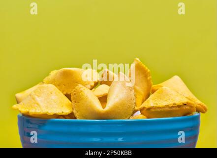 Ungeöffnete Glückskekse in einer blauen Keramikschüssel, teilweise isoliert auf gelbem Hintergrund mit Kopierraum. Stockfoto