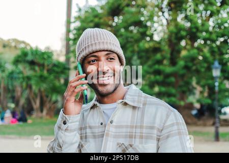 Ein hübscher afroamerikanischer junger Mann, der lächelt und einen Anruf vom Handy hat. Ein glücklicher lateinamerikanischer Mann, der im Park telefoniert. Kommunikationskonzept. Hochwertiges Foto Stockfoto