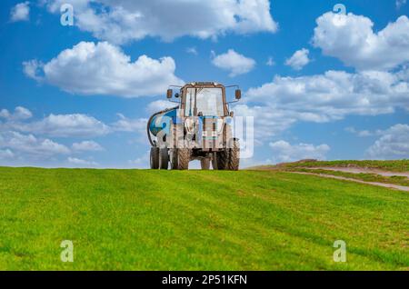 Landwirtschaft, ein Traktor mit einem Fass Anhänger fährt entlang der grünen Feld. Stockfoto