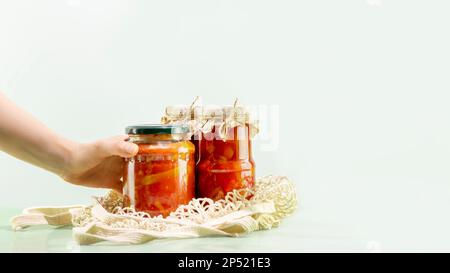 Probiotische Lebensmittel. Nachhaltiges Stillleben mit eingelegtem oder fermentiertem Gemüse mit der Hand einer Frau. Lecho und Ajvar in Glasgefäßen in einem Netzbeutel auf blauem Backgr Stockfoto