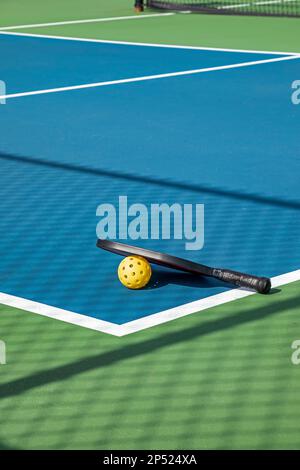 Blick auf eine Pickleball-Anlage mit Paddel und gelbem Ball auf blauen und grünen Plätzen neben einem Spielplatz in einem Vorstadtpark im Frühling. Stockfoto