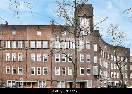 Amsterdam, Niederlande - 10. April 2021: Ein Apartmenthaus mit Autos, die auf der Straße daneben geparkt sind, und einem Uhrenturm in der oberen rechten Ecke Stockfoto