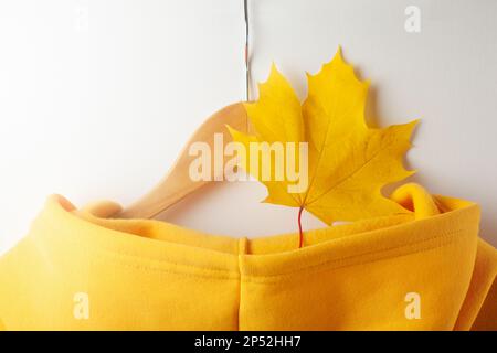 Herbstkonzept - ein gelbes Ahornblatt im Herbst liegt in der Kapuze eines gelben Sweatshirts, das an einem Kleiderbügel hängt. Nahaufnahme Stockfoto