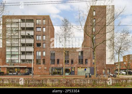 Amsterdam, Niederlande - 10. April 2021: Ein städtisches Gebiet mit hohen Ziegelgebäuden und Bäumen im Vordergrund, von der anderen Straßenseite aus gesehen an einem sonnigen Tag Stockfoto