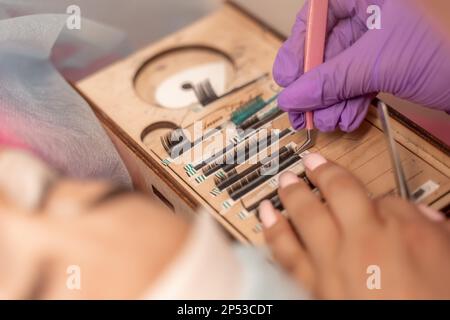 Wimpernverlängerungen, Lashmacher-Werkzeuge. Künstliche Wimpern auf einer weißen Tablette und pinkfarbener Silikonbürstenkamm, Nahaufnahme. Stockfoto