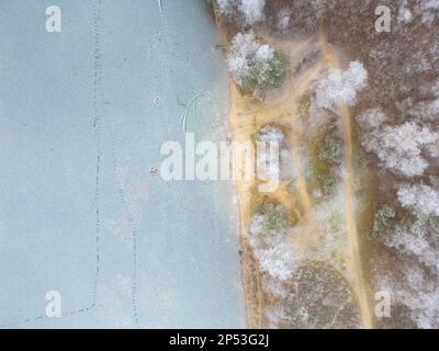 Blick aus der Vogelperspektive oder von oben auf den Winterwaldsee, der das Eis auf dem See und die Kiefern mit schneebedeckten Bäumen zeigt. Winterhintergrundaufnahme durch eine Drohne. Hochwertiges Foto Stockfoto