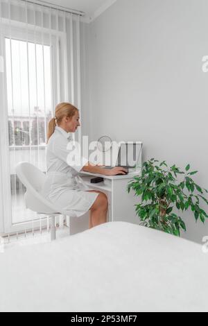 Blonde Ärztin in der Arztpraxis, lächelnd. Auf dem Tisch ist ein Computer, ein Spiegel. Das Büro hat weiße Wände, eine Blume mit grünen Blättern. Stockfoto