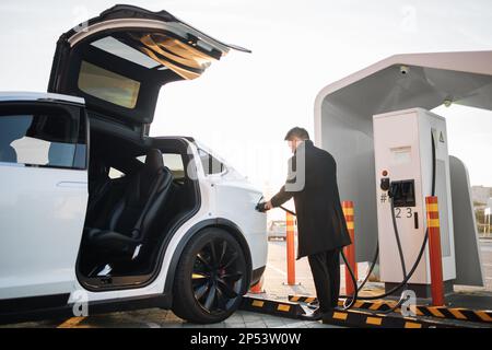 Rückansicht eines stilvollen Geschäftsmanns, der das Netzkabel an sein weißes, luxuriöses Elektroauto an der Ladestation im Freien anschließt. Verkehrskonzept, Ökologie und moderner Lebensstil. Stockfoto