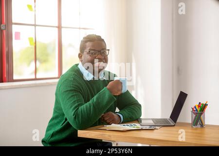 Erfolgreicher Schwarzer Männlicher Designer Mit Laptop, Der Kaffee Am Arbeitsplatz Genießt Stockfoto