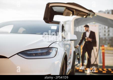 Verschwommener Hintergrund eines indianers in formeller Kleidung, der die Batterie seines Elektroautos an der EV-Ladestation wiederherstellt. Konzentrieren Sie sich auf den weißen, glänzenden Luxus-Auto. Umweltfreundlicher Verkehr. Stockfoto