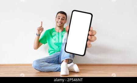Coole App. Happy Arab Guy Sitzt Auf Dem Boden Und Zeigt Ein Leeres Smartphone Stockfoto