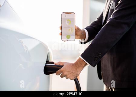 Ernte eines Mannes in schwarzem Anzug, der das Netzkabel einsteckt und den mobilen Bildschirm mit einer App des Ladestatus der Batterie des Elektroautos zeigt. Technologie- und Innovationskonzept. Stockfoto