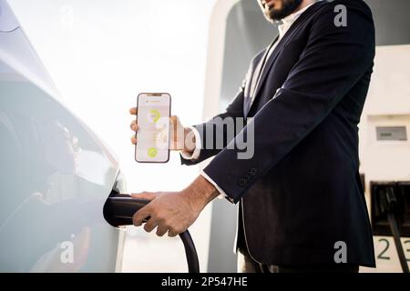 Ernte eines Mannes in schwarzem Anzug, der das Netzkabel einsteckt und den mobilen Bildschirm mit einer App des Ladestatus der Batterie des Elektroautos zeigt. Technologie- und Innovationskonzept. Stockfoto