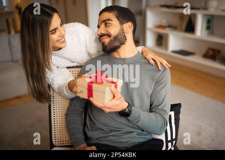 Glückliche junge Weiße gratuliert dem Mann aus dem Nahen Osten, schenkt ihm eine Box mit Geschenk im Zimmer Stockfoto
