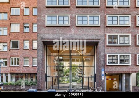 Amsterdam, Niederlande - 10. April 2021: Ein Fahrrad parkt vor einem großen Ziegelgebäude mit vielen Fenstern auf jeder Seite und einem Eingang zur Straße Stockfoto