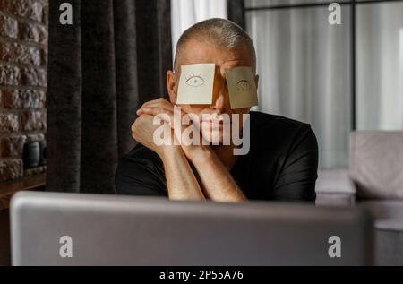 Ein müder Geschäftsmann mit Aufklebern im Gesicht sitzt an einem Tisch im Büro und schläft, die Augen werden auf die Aufkleber gezeichnet. Stockfoto