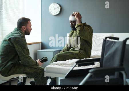 Junger afroamerikanischer Soldat berührte seinen verletzten Kopf gefesselt mit Verband, während er auf dem Bett auf der Krankenhausstation saß und mit einem Freund sprach Stockfoto