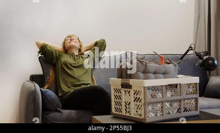 Glückliche Frau, die sich auf der Couch ausruhte, nachdem sie eingezogen war, und eine Kiste auf dem Tisch, zufriedene Hausbesitzerin, die ihren ersten Tag im neuen Haus genossen hat. Hochwertiges Foto Stockfoto