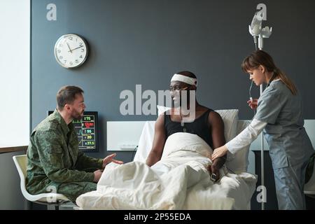 Ein junger Soldat in Militäruniform, der mit seinem verletzten Freund spricht, mit einem Verband, der auf dem Bett sitzt und den Kumpel mit einem Lächeln ansieht Stockfoto