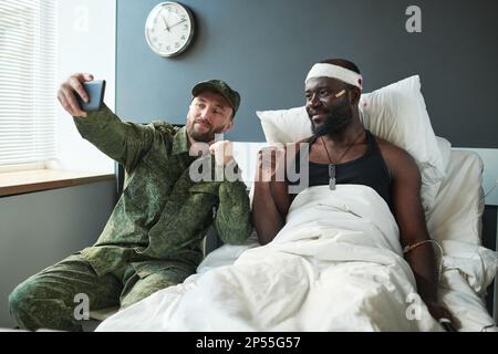 Zwei junge interkulturelle Soldaten, die Selfie auf der Krankenhausstation machen, während einer von ihnen am Bett eines verletzten Freundes mit Verbandskopf sitzt Stockfoto