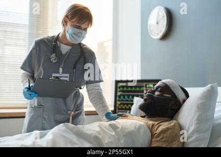 Junge Krankenschwester in Schutzmaske bückt sich über verletzten Soldaten mit Bandkopf schläft im Bett auf der Station des Militärkrankenhauses Stockfoto